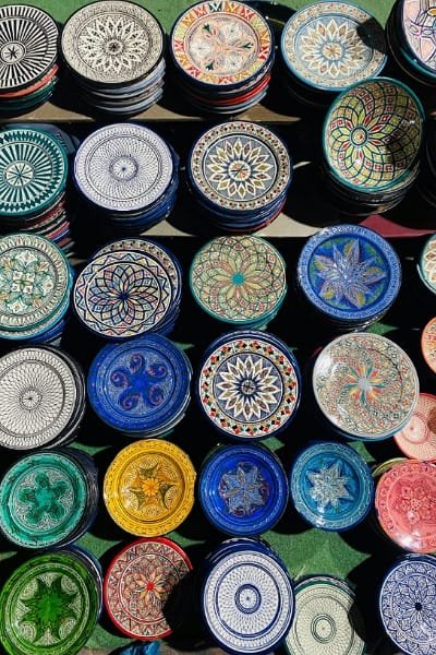 A Moroccan artisan in Fez hand-painting intricate geometric patterns onto a large piece of decorative pottery (often used as a tagine or vase). The work highlights the craftsmanship of Fez hand-painted tagine pots, known for their vivid colors and traditional patterns.