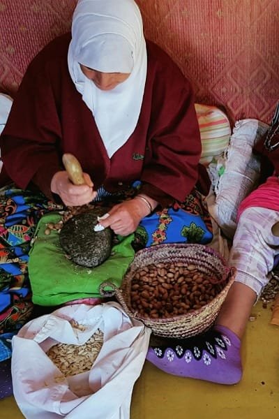 A Moroccan woman in traditional attire manually crushing argan kernels with a stone to extract argan oil. This traditional process highlights the natural preparation of "liquid gold" argan oil, a key ingredient in Moroccan beauty products.