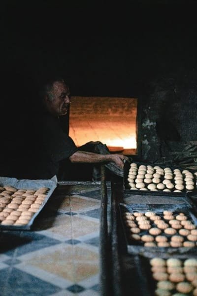 A Moroccan artisan (baker) removing trays of traditional sweets from a warmly lit traditional communal oven. This image reflects ancient Moroccan traditions, simple living, and local heritage.