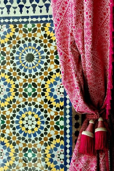 A close-up view of a vibrant, intricate Moroccan Zellige mosaic tile pattern. The hand-cut small glazed tiles form complex geometric designs, reflecting the mathematical precision and artistic heritage of Fez craftsmanship.