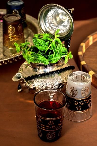 A close-up of a decorative silver Moroccan teapot filled with vibrant fresh spearmint leaves. This image showcases the essential ingredients for authentic Moroccan mint tea and the ornate, traditional presentation with decorated glasses.