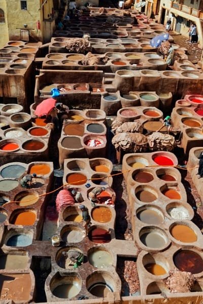 A high-angle panoramic view of the Chouara Tannery in Fez medina, showcasing the numerous dye pits filled with colorful liquids and raw hides. Artisans work diligently below in the world's oldest tannery, symbolizing Morocco's enduring, thousand-year-old traditional leather craftsmanship.