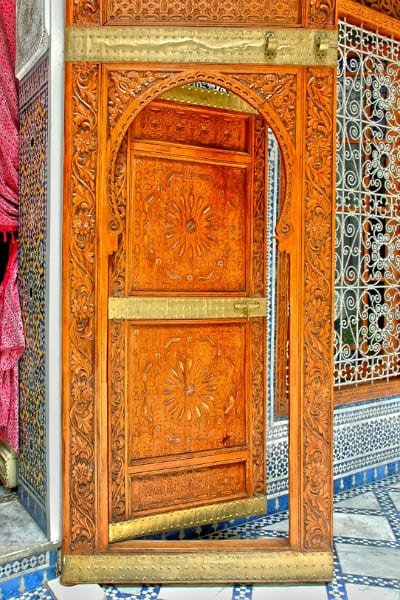 A stunning traditional Moroccan arch doorway intricately hand-carved with arabesque and geometric motifs. The wooden door is adorned with brass accents, flanked by colorful Zellige tiles, symbolizing the spiritual harmony and deep aesthetics of Islamic art in Fez.
