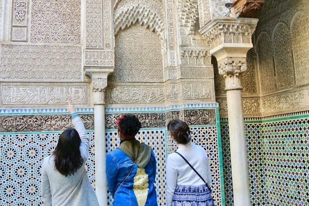 Three tourists inside a Fes historical madrasa, closely examining intricate Moroccan architectural details, including colorful Zellige mosaic patterns and carved Islamic calligraphy on the wall. This image promotes an in-depth private cultural tour and architectural discovery.