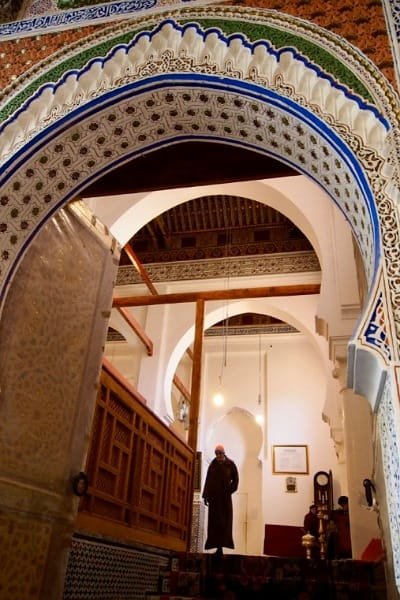 A group of men in traditional Moroccan attire gathered in a deeply shadowed, arched interior, symbolizing the spiritual significance of Moulay Idriss Zerhoun, the holy city and final resting place of Moulay Idriss I, founder of the first Moroccan state. The image reflects the town’s role as a pilgrimage site and center for deep Moroccan history.