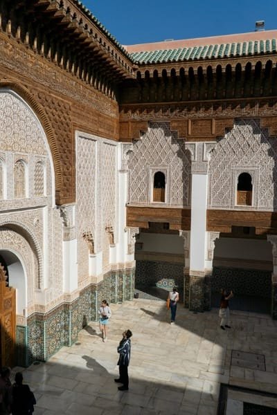 The stunning interior courtyard of Al-Attarine Madrasa in Fes, showcasing exceptional Marinid architecture from 1323-1325. Features include carved cedar wood, white stucco, and vibrant Zellige tiles, emphasizing the harmonious design and spiritual atmosphere of this historic theological school.