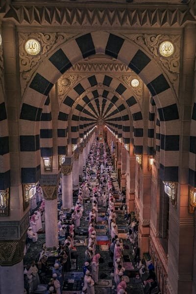 Panoramic view of the Al-Masjid an-Nabawi (Prophet's Mosque) in Medina, featuring long rows of Muslim worshippers praying under striped Islamic arches. This image illustrates the immense scale of the holy site and the spiritual atmosphere, relevant to explaining access rules and sacred boundaries.