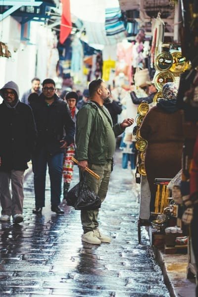 A bustling scene in a narrow Fez souk alley, featuring visitors exploring shops and wet pavement, indicating mild weather (Autumn or Spring). This visual promotes the best time to visit Fez for cultural events and comfortable exploration.