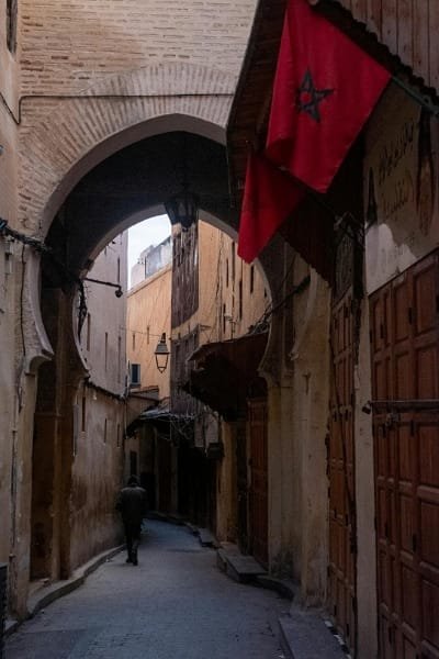 A narrow, arched alleyway (medina labyrinth) in Fez with a person walking, framed by the traditional architecture and red Moroccan flags. The image represents the magical travel experience offered by Fez's souks and traditions, emphasizing the importance of choosing the best time (weather) to visit.