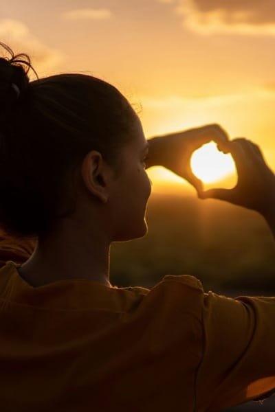 A person making a heart shape with their hands against a golden sunset or sunrise in Morocco. The image beautifully represents the love for Morocco's natural and diverse environment and promotes year-round tourism to enjoy its ecological and biological beauty.