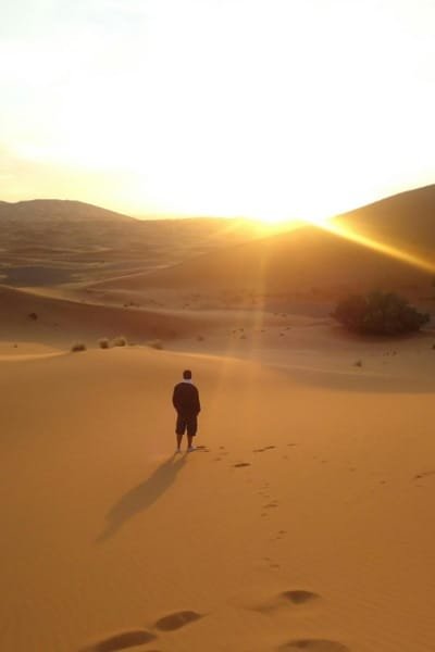 A stunning panoramic view of the Sahara desert dunes during sunset, highlighting the golden light of a summer travel journey in Morocco. A person stands silhouetted on the crest of a dune, capturing the beautiful and vast landscape of the Moroccan desert.