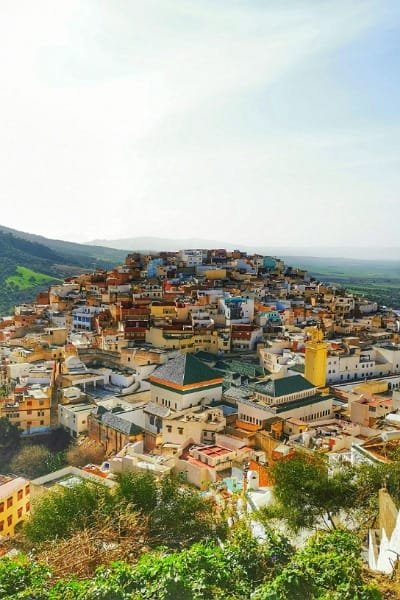 A breathtaking view of the densely packed historic city (Meknes/Moulay Idriss), nestled on a hillside with colorful buildings, traditional roofs, and a mosque minaret. Represents day trips and cultural exploration tours from Fes to nearby imperial or holy cities.
