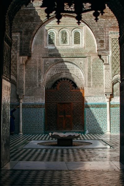 The ornate central courtyard entrance of Al-Attarine Madrasa in Fes, featuring intricate wooden doors, detailed Zellij mosaic tiles, carved stucco, and a central fountain, showcasing the pinnacle of Moroccan Marinid architectural style.