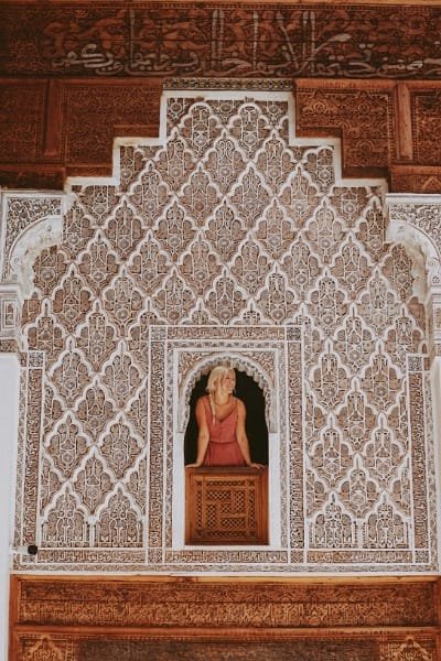 The richly decorated interior of Bou Inania Madrasa in Fes, featuring intricate stucco carving, Zellij, and a visitor looking out from an ornate window. Symbolizes the depth of Moroccan history and architecture.