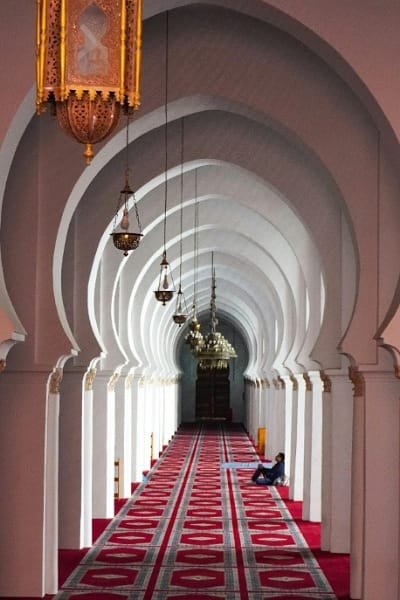 A long hallway in a Moroccan mosque or Riad, featuring repetitive white arches, red carpet, and traditional brass lamps. Symbolizes the profound influence of Islamic art, architecture, and religious pursuits in Morocco since the seventh century.