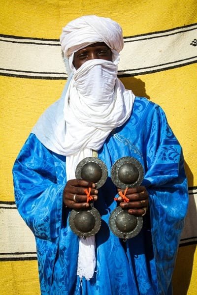 A Gnawa musician in traditional blue robes and turban holding metal castanets. Represents Morocco's deep historical connection to sub-Saharan Africa, evident in the mesmerizing rhythms of Gnawa music.