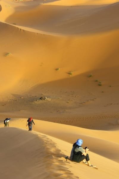 Travelers trekking across the vast, golden Erg Chebbi dunes near Merzouga in the Moroccan Sahara Desert. Represents a transformative journey into infinite horizons with camel rides and spending the night under a million stars in a traditional Bedouin camp.