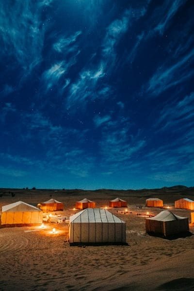 A stunning night photograph of a luxury Bedouin camp set among the sand dunes, with illuminated tents and a vibrant, starry night sky overhead. Represents an unforgettable night stay in the Sahara Desert, offering a truly transformative experience.
