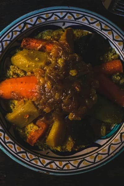 A close-up of the Moroccan national dish, Couscous, served in a traditional, intricately hand-painted platter. The dish features tender meat (hidden), fluffy semolina grains, and a vibrant assortment of steamed vegetables (carrots, zucchini, squash).
