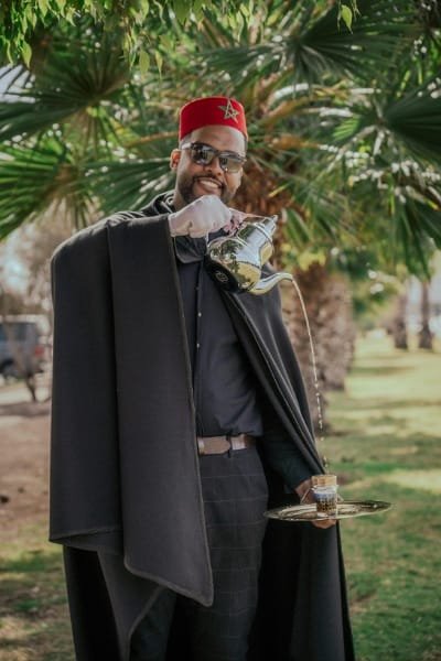 A man in a traditional Moroccan Selham and Tarboosh pouring fresh mint tea into a small glass on a metal tray. Represents the famous Moroccan Mint Tea, a profound symbol of hospitality and tradition, often dubbed 'Berber whiskey'.