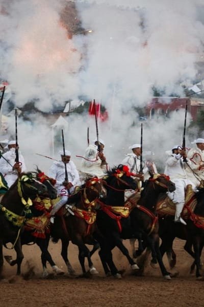 A friendly local guide (representing the values of Joud/generosity) standing with a tourist couple in a richly decorated doorway of a Fes Riad or shop entrance. This interaction embodies the profound Moroccan hospitality rooted in Islamic, Arab, and Amazigh values, ensuring guests feel comfortable and safe.