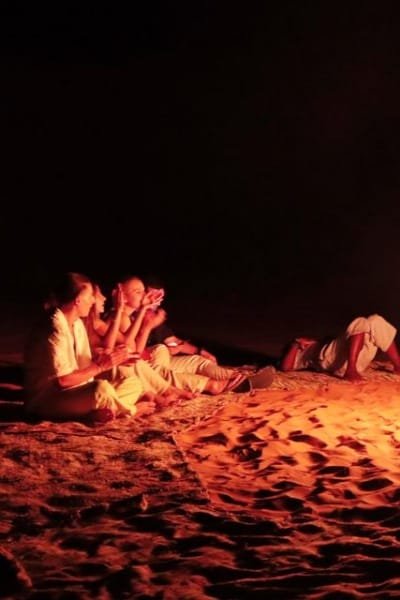 A group of travelers sitting together on the ground in a warm, low-light setting (possibly a desert camp or a night gathering). This image represents the singular human experience based on authentic Moroccan hospitality, where every visitor is treated as a valued member of the family.