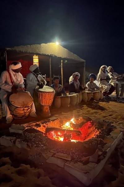 A group of local musicians (Berbers/Tuareg) playing drums around a blazing campfire at night in a Moroccan luxury desert camp. This scene represents the authentic Moroccan hospitality and entertainment, including singing, music, and dance activities.