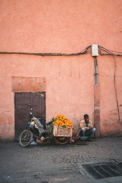 A man rests next to a motorbike laden with oranges against a warm, pink wall in a Moroccan city (likely Marrakech). This street scene represents the urban environment where "petite taxis" are readily available for affordable, short-distance travel.