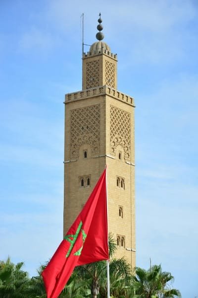 A large, towering minaret (representing a religious site or local landmark) with the Moroccan flag flying in the foreground. Symbolizes the conservative, Islamic culture of Morocco, emphasizing the importance of dressing modestly and being mindful of local customs to ensure a safe and respectful experience.