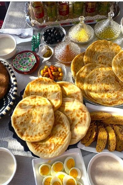 A rich traditional Moroccan breakfast table featuring Moroccan breads (Khobz/Batbout), boiled eggs, local olives, honey, and glasses of mint tea. This image highlights the hospitality and diverse, hearty nature of Moroccan cuisine.