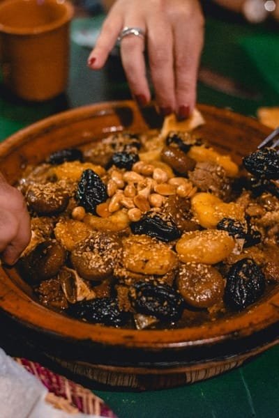 A group of people sharing a traditional Moroccan tagine (likely beef with prunes and almonds) served in an earthenware pot, showcasing the authentic flavors and communal dining style of Morocco. This dish is the centerpiece of a hearty, flavorful meal enjoyed with friends.