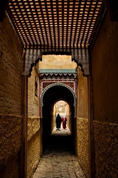 A narrow, covered alley (derb) in the Fez medina, featuring traditional archways, carved wooden ceiling, and intricate tilework. This area is representative of the quiet, private residential life that thrives away from the main tourist paths.