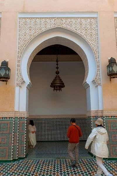 Tourists navigating a beautiful, complex courtyard or madrasa in the Fez medina, highlighting intricate Moroccan zellige tiles and arches. This illustrates the value of a local guide in Fez for understanding the history behind the 9,000 narrow alleys and avoiding confusion.