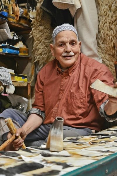 A smiling elderly artisan sitting in his workshop in Fez, surrounded by traditional handcrafted goods and tools. This image represents Fez as the craft capital of Morocco and highlights the authentic shopping experience in the medina.