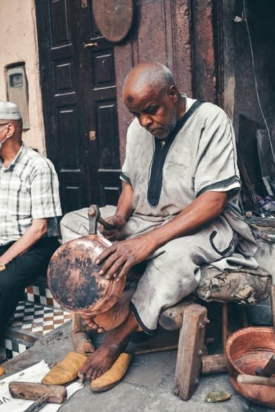 A skilled copperware artisan in Fes el Bali hammering and shaping a metal pot using ancient, preserved techniques. The image highlights the detailed craftsmanship involved in creating Moroccan copper pots and trays.