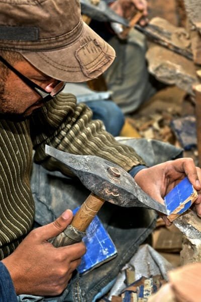 A close-up of a master artisan engraving a colorful zellij tile using a hammer and chisel. This process creates the intricate geometric mosaics that define Moroccan architecture in Fez's palaces, mosques, and fountains.