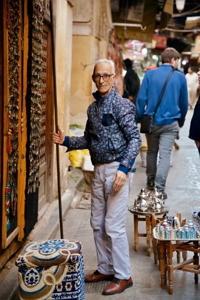A man in traditional Moroccan attire (Jellaba and Fez) pouring mint tea high into a small glass on a tray, symbolizing the genuine Moroccan ritual of sharing tea and tales with locals, known for their warmth and generosity.