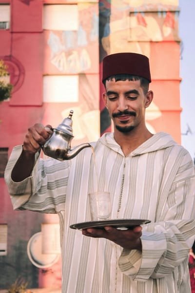 A Moroccan man in traditional attire (Jellaba and Fez) meticulously pouring mint tea from a silver teapot into a glass with a characteristic high flourish. This image highlights the deep-rooted cultural ritual of hospitality, friendship, and the sweet, fragrant aroma of fresh mint tea.