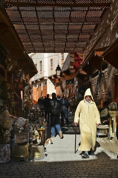 A covered, specialized alley (souk) in Fez, likely the copperware souk, with traditional metal goods displayed alongside a local man in a Jellaba. The image represents the segmented Fez markets, such as Souk Attarine for spices or Souk Sabbaghine for textiles.
