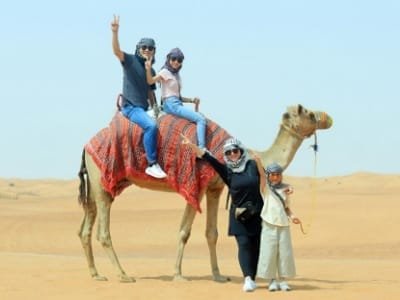 A joyful family (parents and children) posing on and next to a camel in the vast golden Sahara Desert, promoting the 3-day luxury tour from Fes to Merzouga and Ouarzazate.