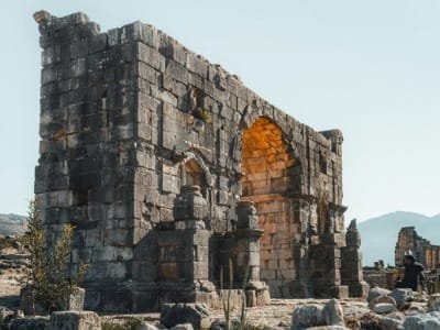 Ancient stone ruins of the Roman city of Volubilis, promoting a historical day trip tour from Fes to Volubilis, Moulay Idriss, and Meknes, Morocco.