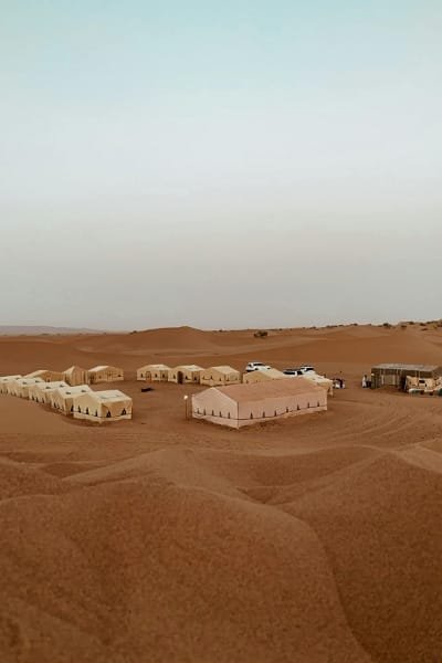 Panoramic view of a full luxury desert camp with multiple tents set up in the vast sand dunes of the Merzouga Sahara, promoting unique overnight stays.