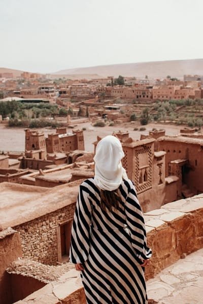 Tourist standing and looking at the panoramic view of the historic Kasbah in the Ouarzazate region (Aït Benhaddou), promoting tours to explore historical and heritage sites.