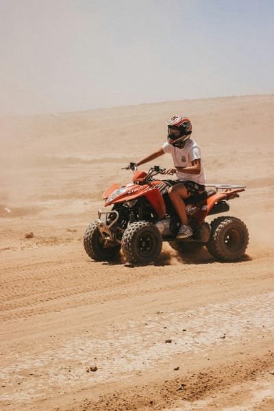 Young person wearing a helmet riding a red quad bike (ATV) on a dusty desert track under the bright sun.