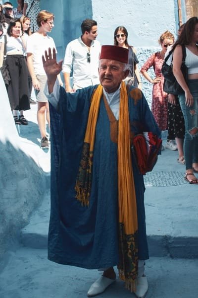 A friendly Moroccan man in traditional blue clothing and a red Fez hat waves in the narrow, blue-painted streets of Chefchaouen, promoting a cultural day trip with scenic stops.
