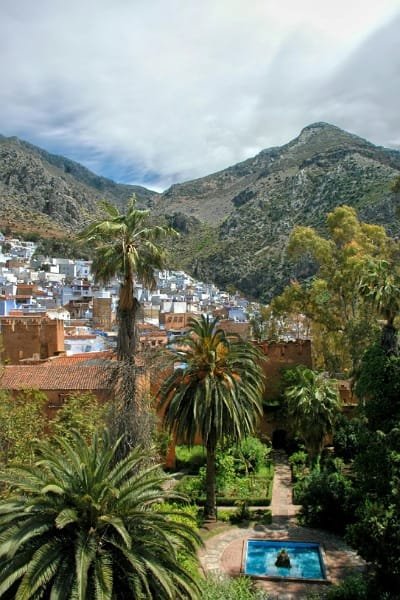 A panoramic view of the blue city of Chefchaouen nestled beneath the Rif Mountains, featuring green palm gardens and a blue pool in the foreground, reflecting the city's natural beauty.