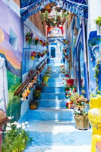 Narrow, blue stairs in a Chefchaouen alleyway decorated with colorful flower pots, small pom-pom banners, and wall murals, highlighting the city's artistic and aesthetic appeal.