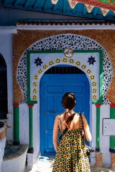 A tourist stands facing an ornate blue Moroccan door with traditional mosaic and arches in Chefchaouen, highlighting the city's unique architecture and culture.