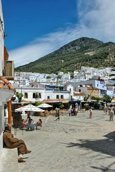 A view of the public square (likely Place Outa Hammam) in Chefchaouen, where locals and tourists sit at cafes under white umbrellas, with a background of a towering mountain and white and blue houses.