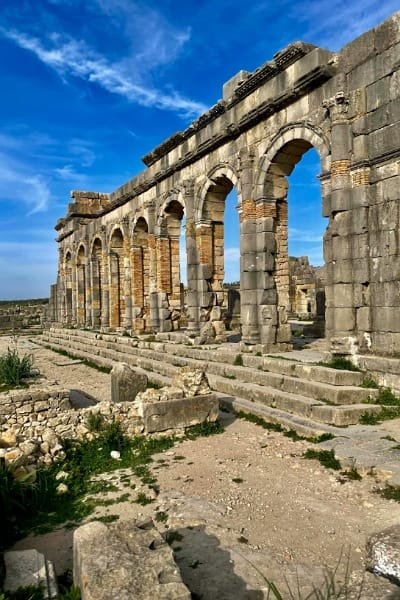 The Roman Triumphal Arch of Caracalla at the Volubilis archaeological site, surrounded by vast green fields and blue sky, representing the major landmark on the historical day trip from Fes.